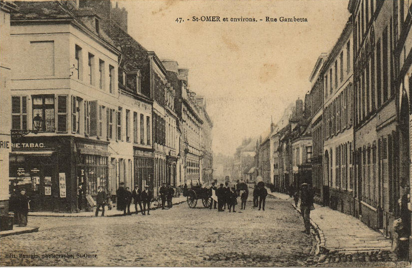 On peut voir à ce carrefour avec la rue du Lycée, une épicerie-tabac, ce qui était trés fréquent à l'époque. Les pavés qui recouvraient la chaussée de nombreuses rues de Saint-Omer sont bien visibles sur cette carte. Si vous vous baladez aujourd'hui dans ce lieu, vous verrez qu'ils sont encore présents au sol.